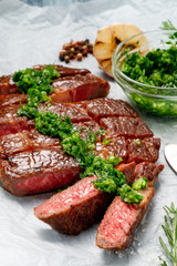Sliced grilled rib eye beef steak with chimichurri sauce on white paper. Concept of cooked steak