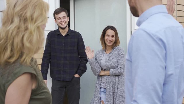 Pleasant caucasian family standing at doors, waving and smiling. Guests leaving their new neighbours. Married couples saying goodbye to each other.