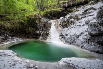 Wide angle shot of an alpine landscape with a waterfall flowing between white rocks and into an emerald-green pond, surrounded by vegetation