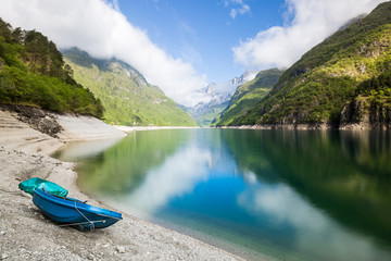 Alpine lake with a beach and a blue wooden boat in the foreground, and sloping mountains and distant clouds in the background, reflecting on the water © Roberto