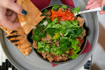 woman serving shakshuka with sausage, tomatoes, egg, parsley, in cast iron skillet with pita bread