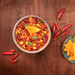 Chili con carne with chilli peppers and nachos, overhead square shot on a dark rustic wooden background