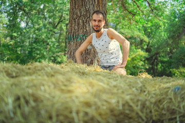 Naklejka na ściany i meble A young handsome man with beard of slim build sits in the hay and holds hay in his hands against the background of green trees in the park.