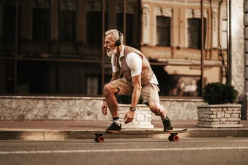 Porträt eines bärtigen Hipster-Mannes in weißem Hemd auf der Stadtstraße. Stilvolle, glückliche Modellfahrt auf Longboard in der Nähe der Straße auf Gebäudehintergrund © Yevhen