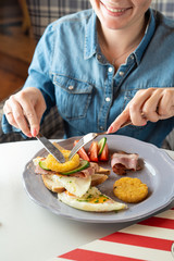 Young woman is eating british breakfast in a cafe: fried egg, wheat crunchy toasts, potato hash browns, bacon and salad