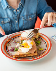 Young woman is cutting a poached egg with knife and fork as a part of breakfast with soft waffle, mashed avocado and salmon