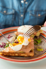 Close up of cutting a poached egg with knife and fork as a part of breakfast with soft waffle, mashed avocado and salmon