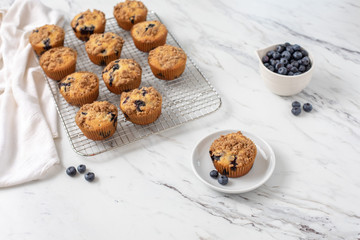 Homemade Blueberry Muffins on Cooling Rack with One Isolated in Front on Small White Plate; Bowl of Blueberries in Background; White Countertop