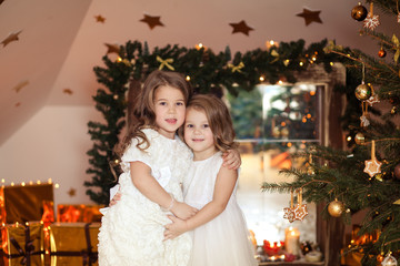 Happy cute little sisters girls in a white dress near the fireplace with candles in a festive atmosphere in anticipation of a Merry Christmas.