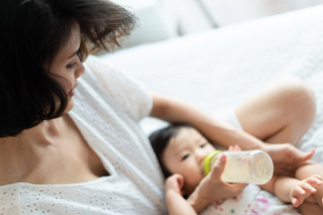 Obraz na płótnie Canvas Pretty Asian mother feeding milk to lovely baby lying on her legs. Mom holding bottle of breast milk helping the child drinking on bed in bedroom. Love, relationship of parent and kid. Health concept.