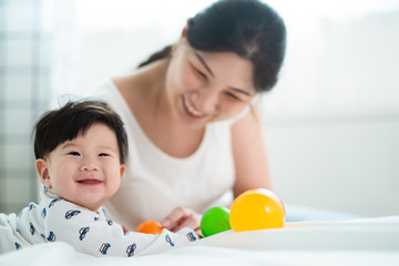 Obraz na płótnie Canvas Asian cute mother and baby playing colorful ball on the bed. Mom touching her kid while the child is smiling with happiness. She looking her son with love and care. Love relationship of parent.