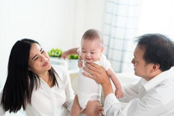 Obraz na płótnie Canvas Asian parents holding and playing with newborn baby in bedroom. Father holding son in hand and Mother supporting the leg and smile to their child. Kid feeling happy with smile to be with family.