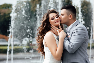 Fototapeta premium Young man kisses his beautiful girlfriend. Romantic, tender moment of a young attractive couple. Happy girl in white dress and guy posing in grey suit. Lovely couple near fountain.