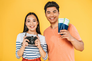 pretty asian girl with digital camera and man with passports and air tickets smiling at camera isolated on yellow