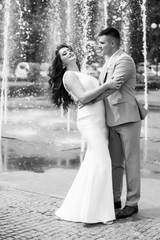 Beautiful smiling love couple danse near fountain outdoors in the city. Beautiful long-haired girl in luxury dress with her handsome husband in grey suit walking . Sweet pair posing.  Black and white