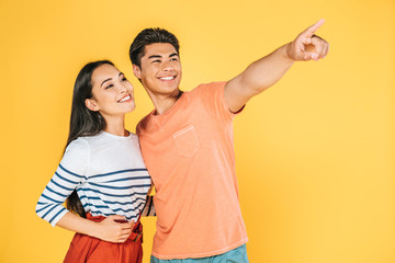 happy asian man looking away and pointing with finger while standing near smiling girlfriend isolated on yellow