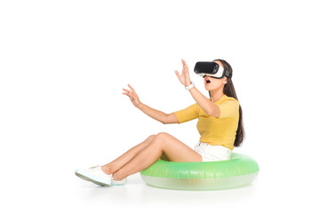 shocked asian woman using virtual reality headset and gesturing while sitting on swim ring on white background