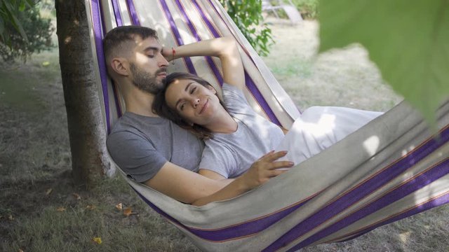 Cute young caucasian man and woman lying in hammock in the garden relaxing together. Loving couple together outdoors. Summertime leisure