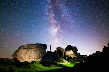Silhouette of figure stargazing in rocky landscape below a clear night sky & vibrant Milky Way,...