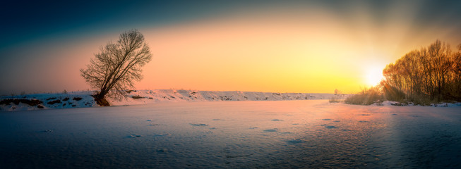 Beautiful winter golden hour panorama with the lonely tree and frozen river after sunset