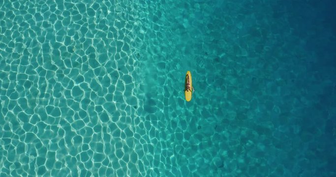 Aerial view of female surfer paddling yellow longboard surfboard in pristine blue ocean lagoon, surf adventure lifestyle