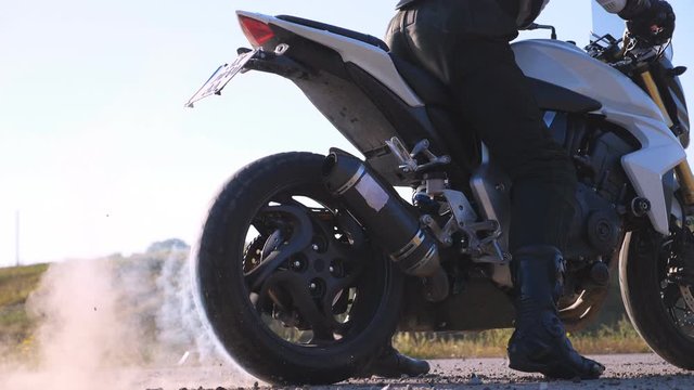 Wheel of sport motorbike starting to spin at asphalt and kicking up pebbles and dirt. Biker performing tire burnout with lots of smoke. Motorcycle starts the movement. Slow motion Close up