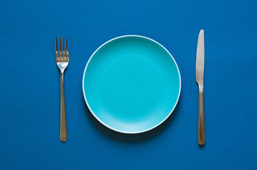 Ready to order. High angle shot of empty color plate, fork, knife lying against blue background.