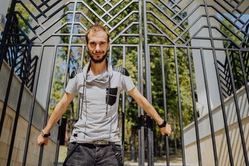 A young handsome man with beard of slim build in casual clothes poses against a black fence on a sunny day.