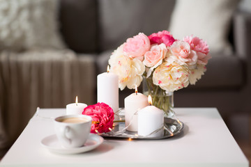 hygge and aromatherapy concept - burning candles, electric garland lights, cup of coffee and flowers on table