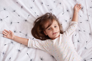 Top view of little girl laying in bed and spreading her arms on white cover with dandelion,...