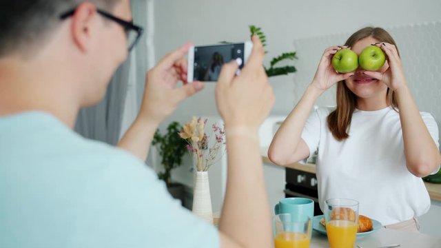 Slow motion of young lady posing with apples having fun when guy is taking photo with smartphone camera touching screen. Youth and photograph concept.