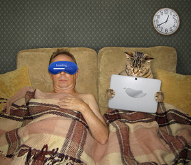 The man in a funny blue sleeping mask and the cat with a laptop are in a bed in the bedroom together.