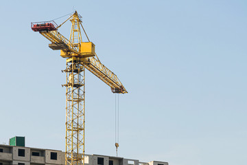 A yellow high-rise building crane against a blue sky builds multi-storey apartment buildings using modern technologies of metal, concrete and brick according to an architectural project