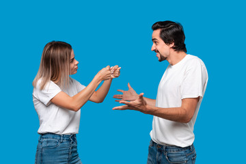 Couple of a young blond woman and brunet bearded man with mustaches in white t-shirts and blue jeans quarreling standing isolated over blue background. Concept of relationship crisis.