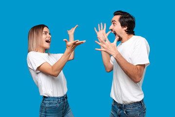 Emotional couple expressing gesture of success. Young blond woman and bearded man with mustaches in white tees and blue jeans are winner. Models standing isolated over blue background.