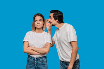 Naklejka premium Bearded man with mustaches in white t-shirt tells secret to a young blond girl in white t-shirts and blue jeans standing isolated over blue background. Concept of secret telling.