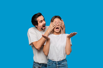 Fototapeta premium Brunet bearded man with mustaches in white t-shirts and blue jeans making a gift for a young blond woman isolated over blue background. Surprise for a girlfriend.