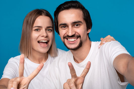 Happy couple of a young blond woman and brunet bearded man with mustaches in white t-shirts and blue jeans making selfie holding thumbs up isolated over blue background. Concept of an ideal couple.