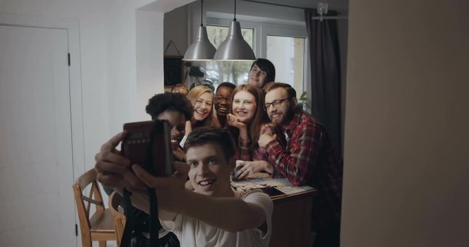 Happy fun young multiethnic friends take an instant selfie group photo sitting together in kitchen at party slow motion.