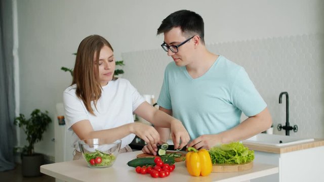 Husband and wife happy young people are cooking salad together cutting fresh raw vegetables in kitchen at home. Lifestyle, nutrition and family concept.