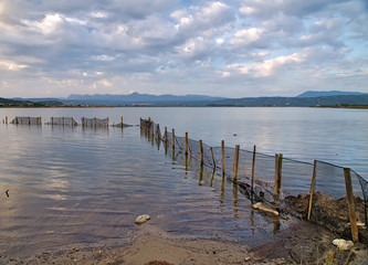 Wooden posts with metal mesh fence in Gialova sea lagoon, Peloponesse, Greece. Dramatic cloudy sky over sea rippled water surface.