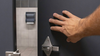 Close up of tan hairy man hand pushing open a grey blue bathroom stall door from inside under a warm light front point of view perspective angle.