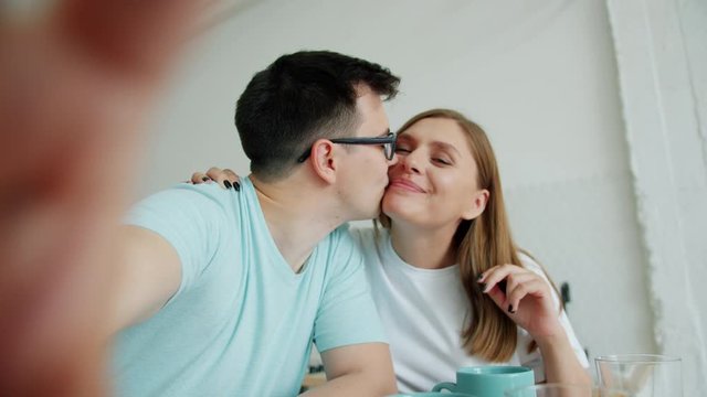 POV portrait of happy young girl and guy kissing having fun taking selfie holding camera in kitchen. Modern technology, relationship and lifestyle concept.