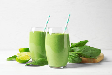 Glasses of healthy green smoothie with fresh spinach on white wooden table against light background