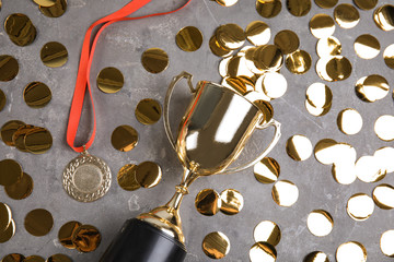 Gold trophy cup, medal and confetti on grey stone background, flat lay