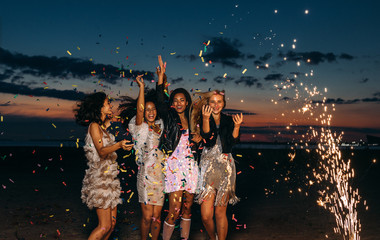 Happy friends celebrating outdoors at sunset. Four stylish women throwing confetti outdoors near fireworks. - obrazy, fototapety, plakaty
