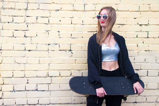 fashion woman with a skateboard stand on brickwall background. Girl in sunglasses with longboard