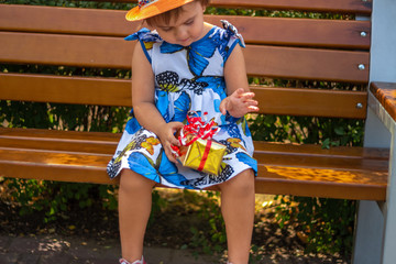 Cute little girl in hat sitting on a bench at the park with a present box. Summer. Close up