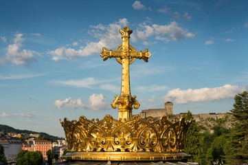 View of the golden cross located in the Catholic sanctuary of Our Lady of Lourdes in the Pyrenees Mountains (France), with Lourdes Castle in the background.