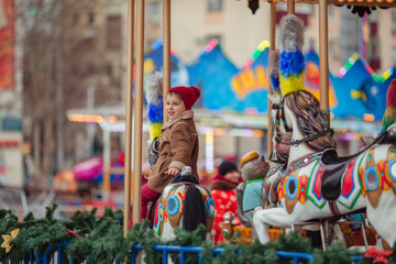 Fototapeta na wymiar Cute smiling boy in a brown coat and a red hat rides on a merry-go-round.